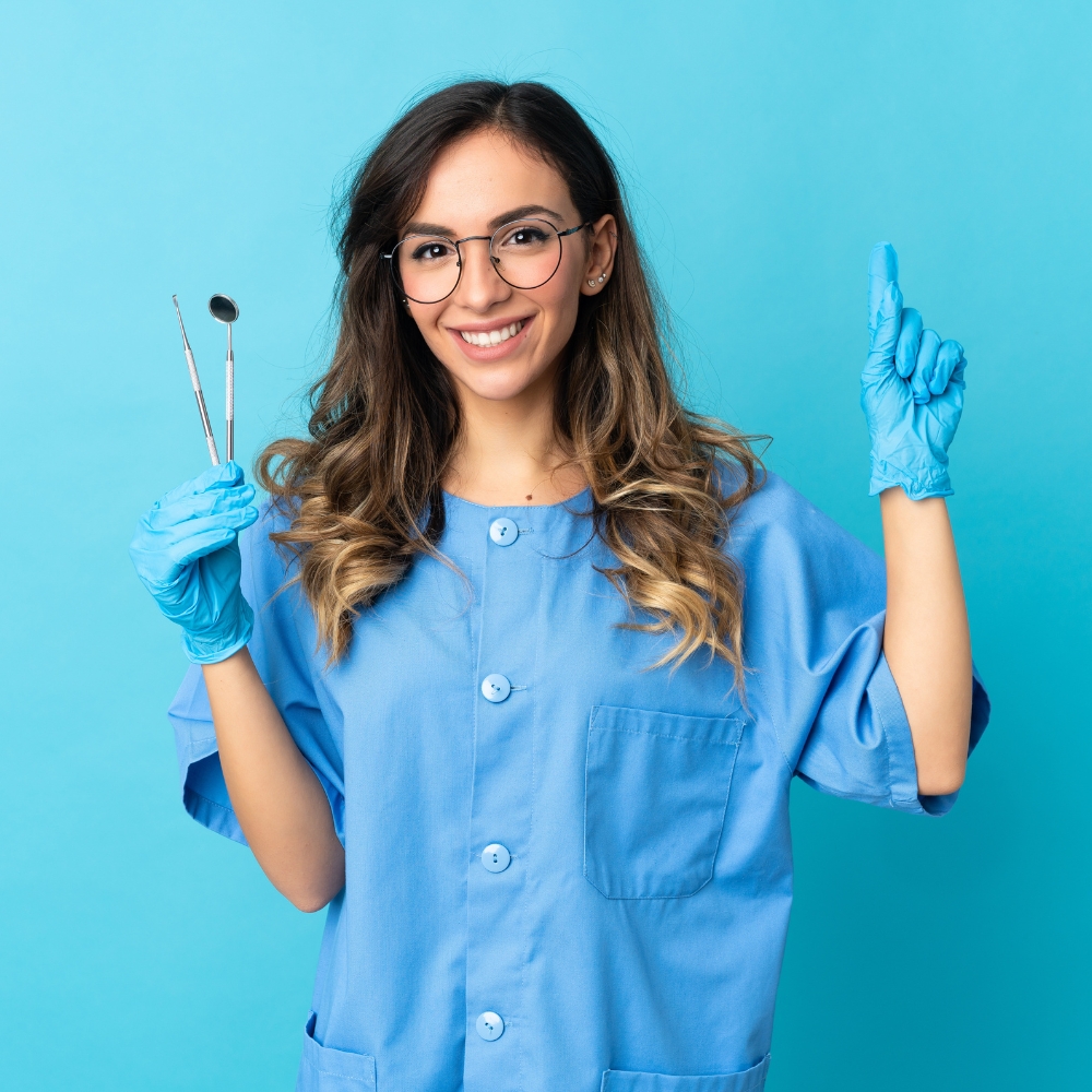 A smiling dental assistant in blue scrubs and gloves holding dental tools in one hand and pointing upwards with the other, against a bright blue background.