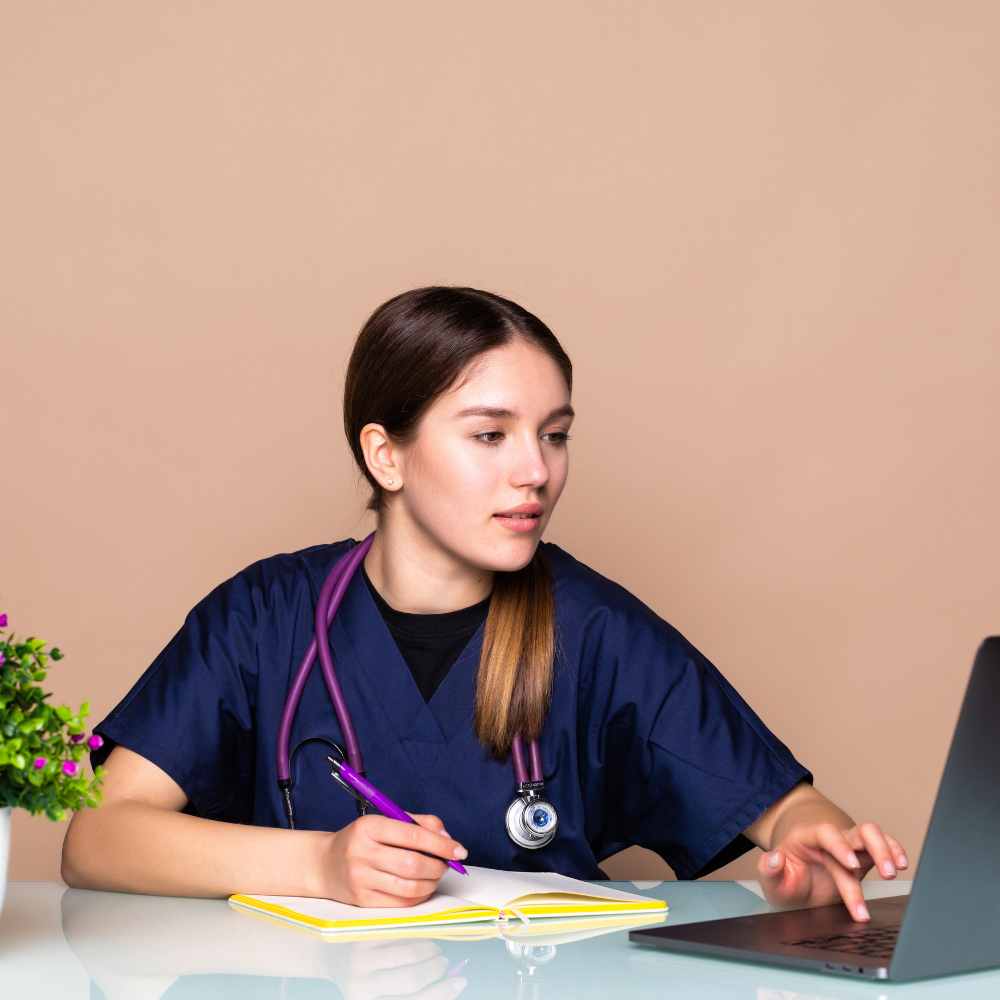 A healthcare professional in navy blue scrubs with a stethoscope, writing in a notebook while working on a laptop at a desk.