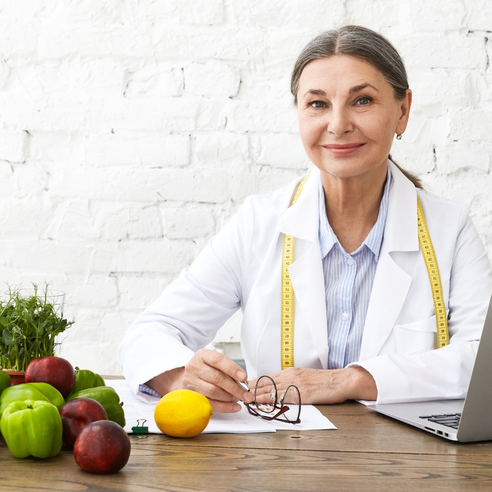 A professional dietitian in a white coat sitting at a desk with fresh fruits, vegetables, and herbs, smiling confidently with a measuring tape around her neck.